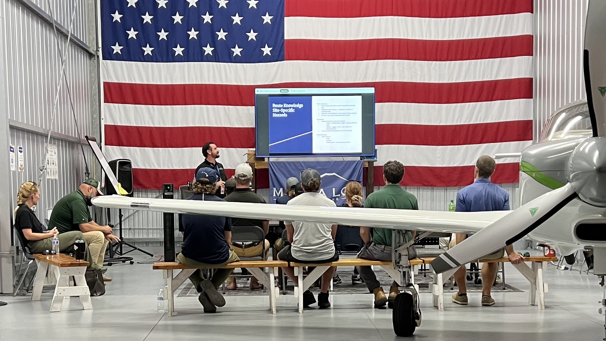 IMG_2474 People seated watching a presentation with a plane and USA flag in the background.