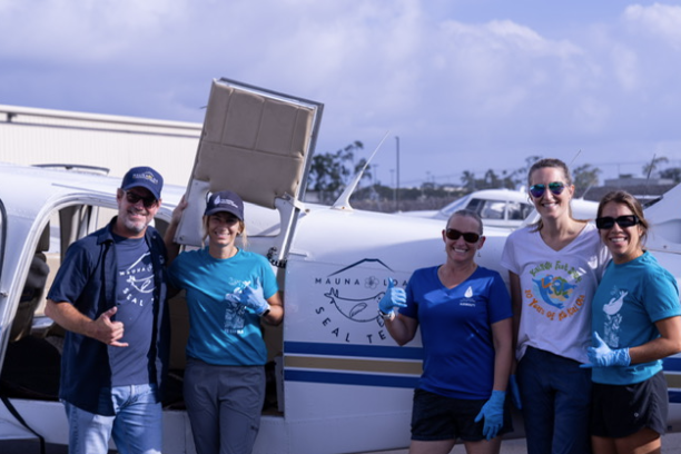 Five people smiling and posing in front of a small airplane on a sunny day.